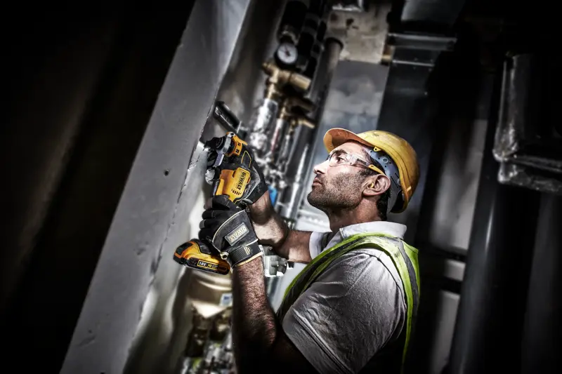Worker uses a DEWALT DCS355D2 power tool on a wall at a construction site, wearing safety gear and gloves.
