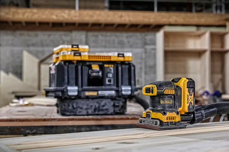 DEWALT cordless sander on wood, with storage box in background in a workshop setting.