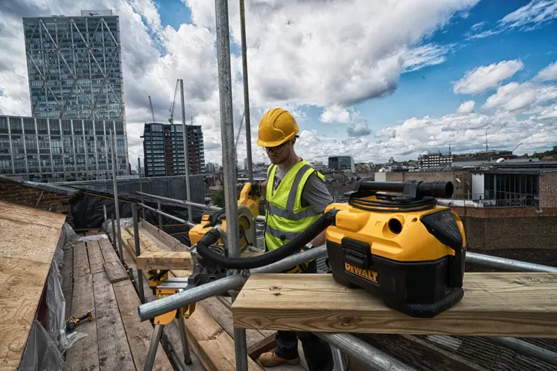 DEWALT DCV584 vacuum on a rooftop construction site with a worker in safety gear and city buildings in the background.