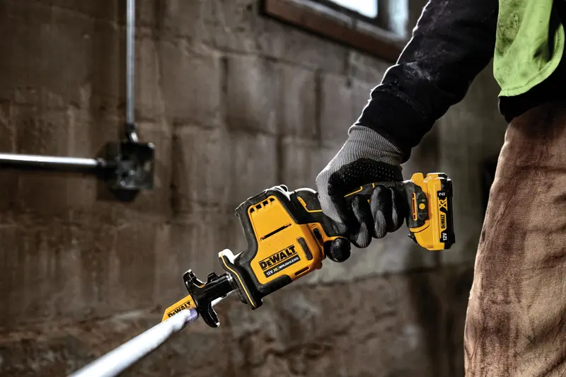 Gloved hand holding a DEWALT DCS312D2 cordless reciprocating saw cutting a metal pipe indoors.